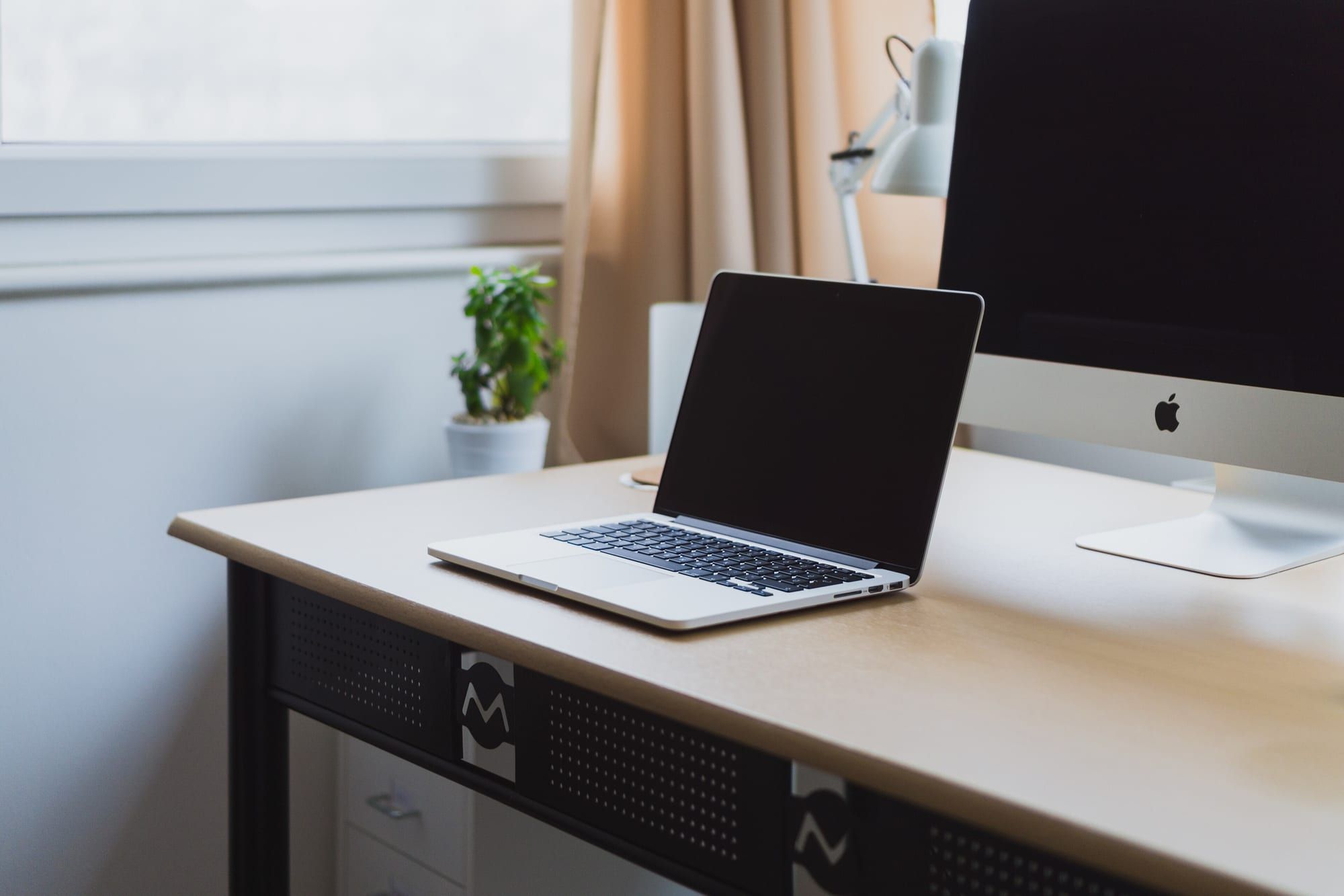A laptop open on a light wooden desk beside a window, with a desk lamp and a houseplant