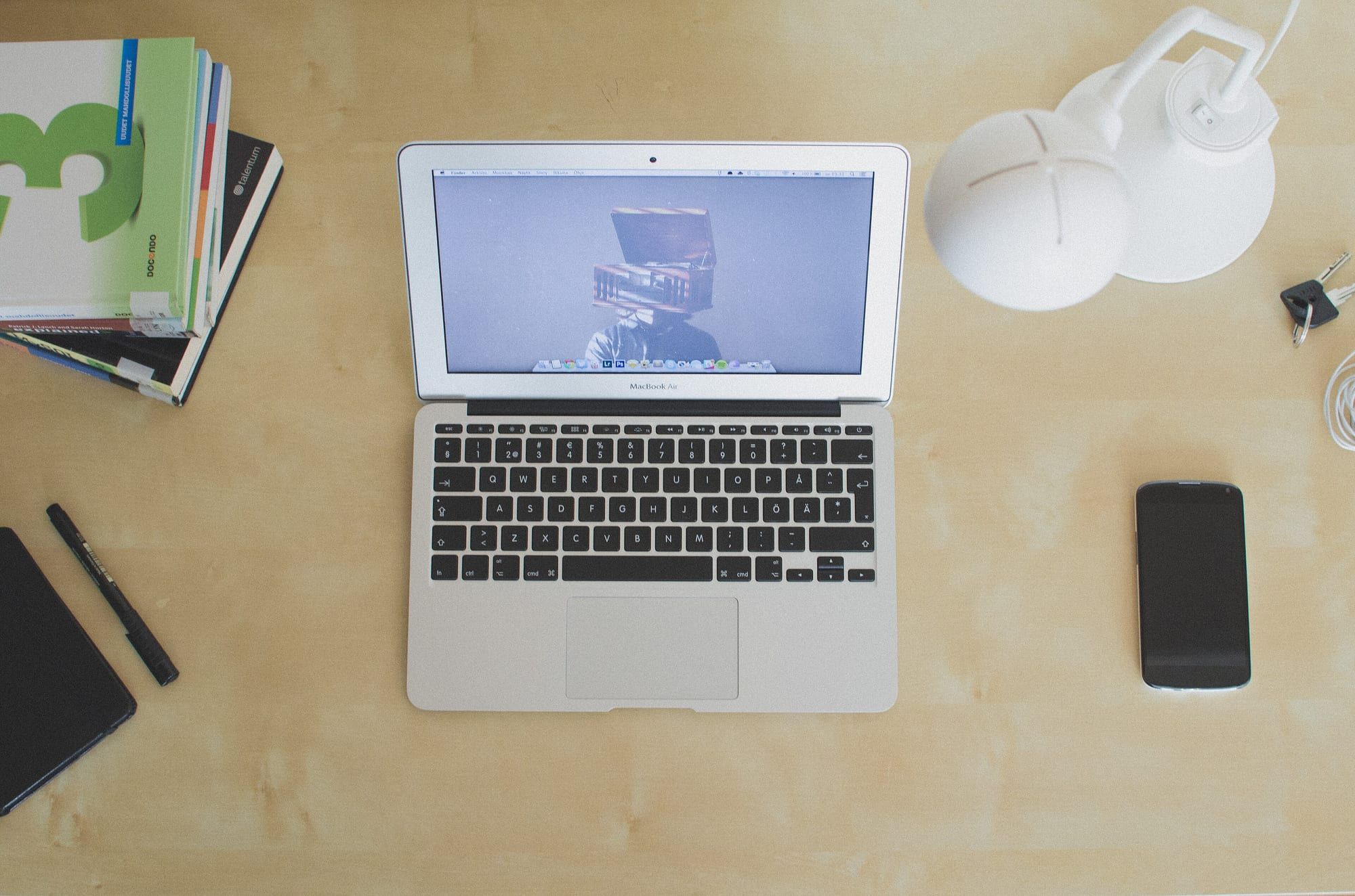 An open silver MacBook Air seen from above on a light wooden desk, with a stack of textbooks, a white desk lamp and a smartphone beside it