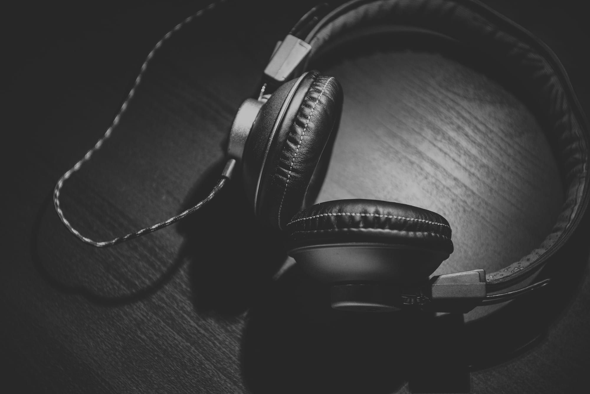 A pair of black over-ear headphones with leather-style ear cups lying on a dark wooden surface, photographed in black and white