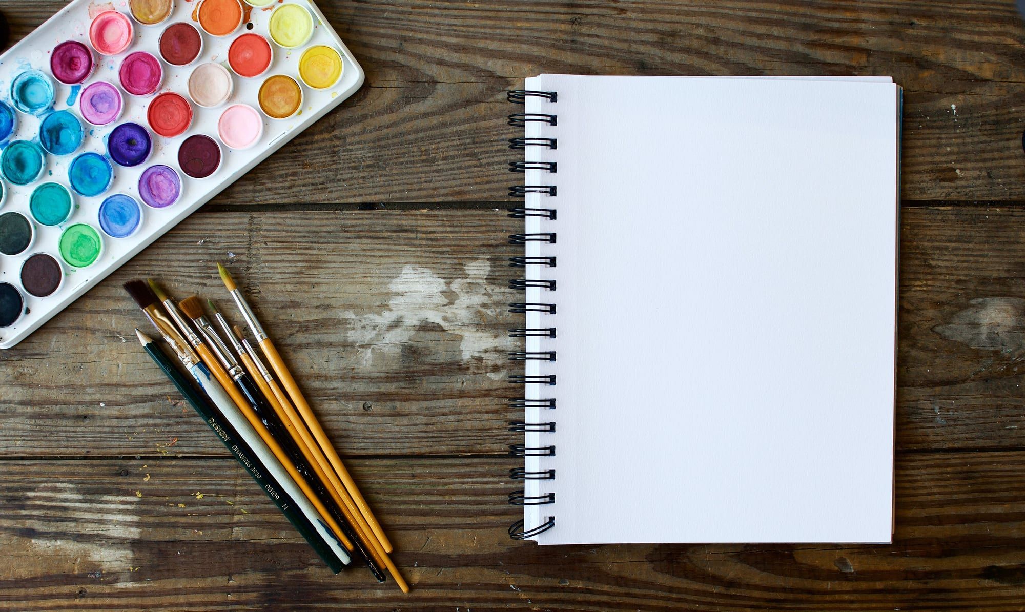 A blank spiral sketchbook beside a watercolour paint set and brushes on a wooden table