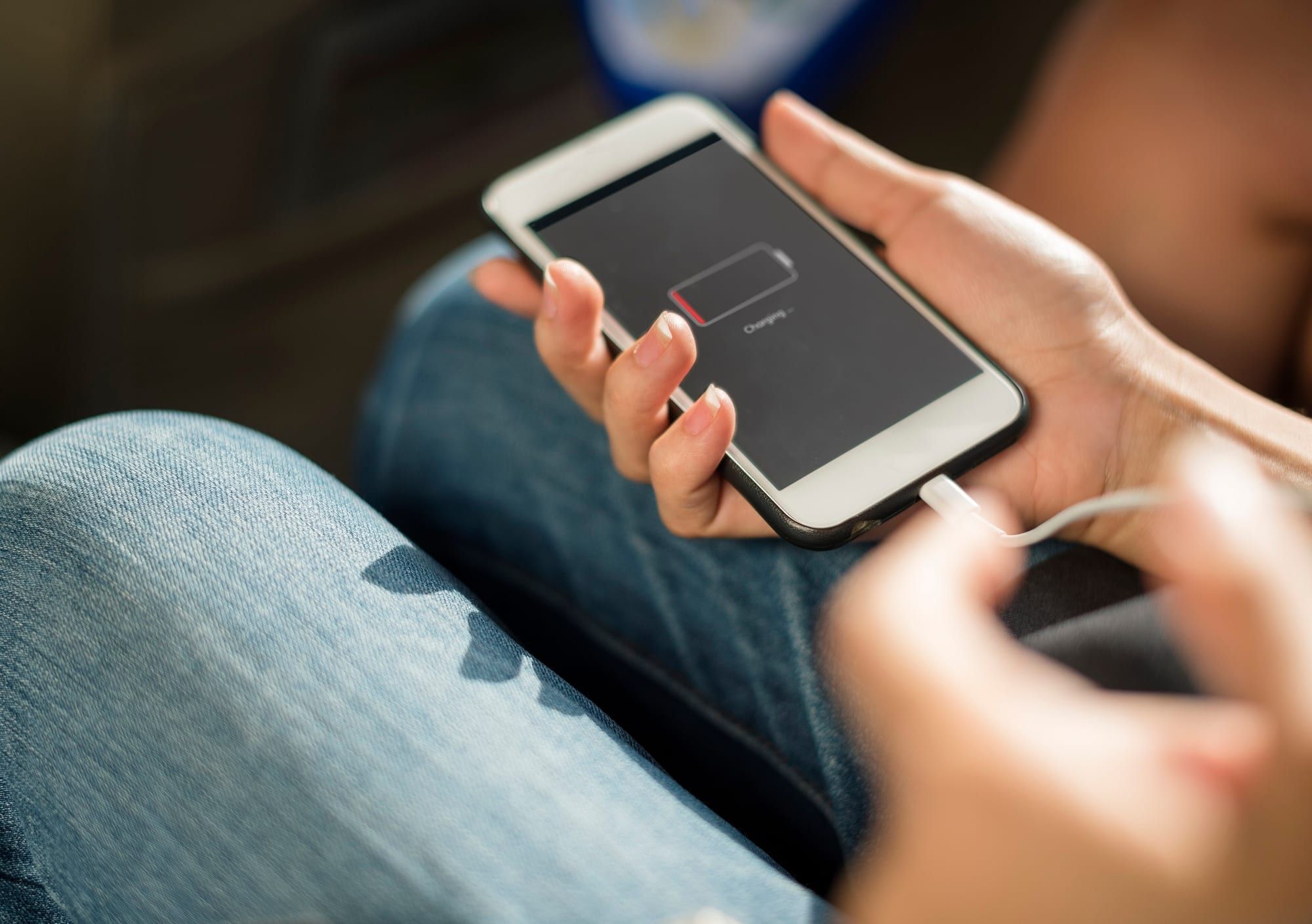 Two hands holding a smartphone that shows a low-battery charging screen, with a charging cable plugged into the phone