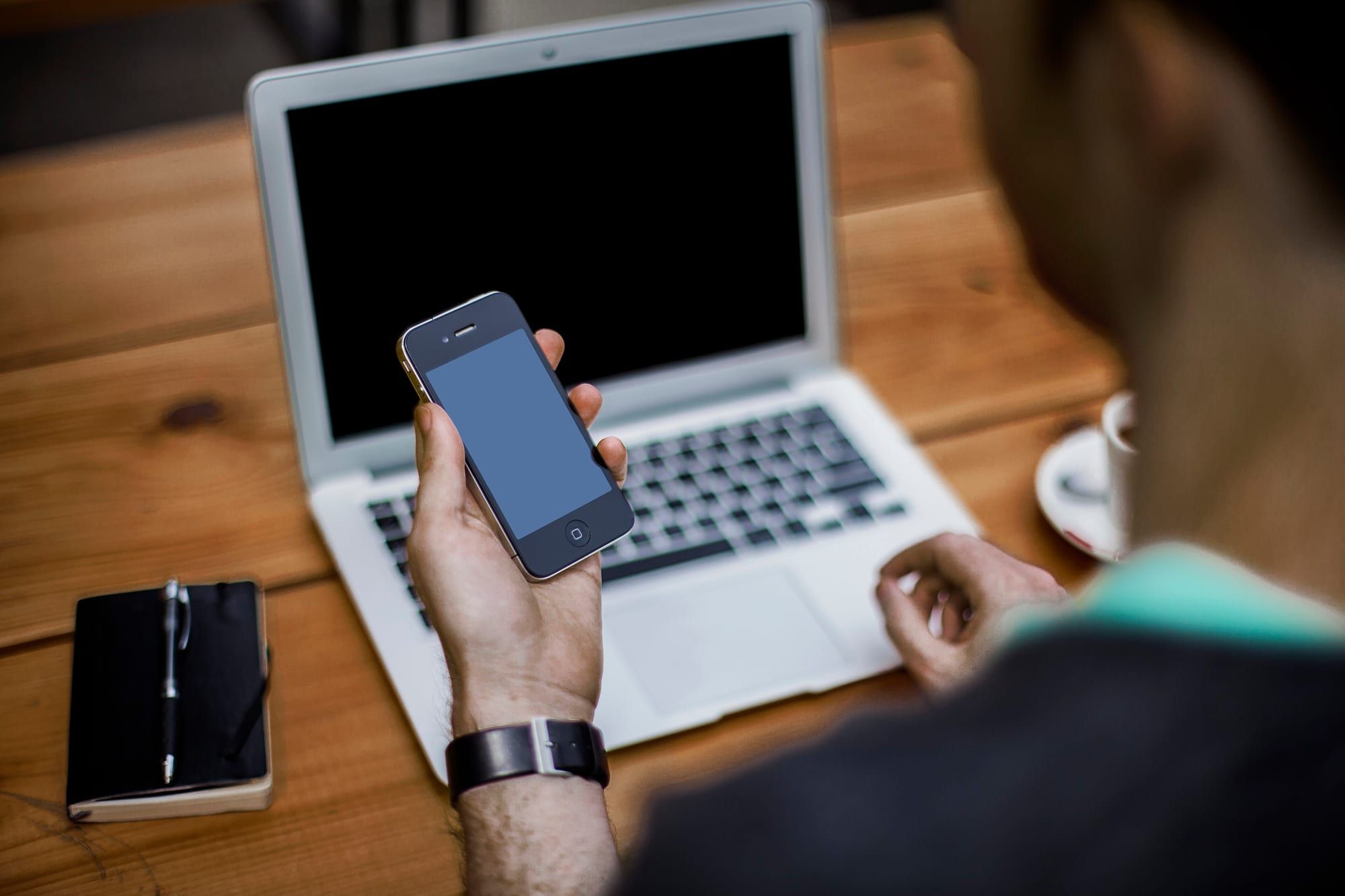 A person holding a smartphone in front of an open laptop on a wooden table