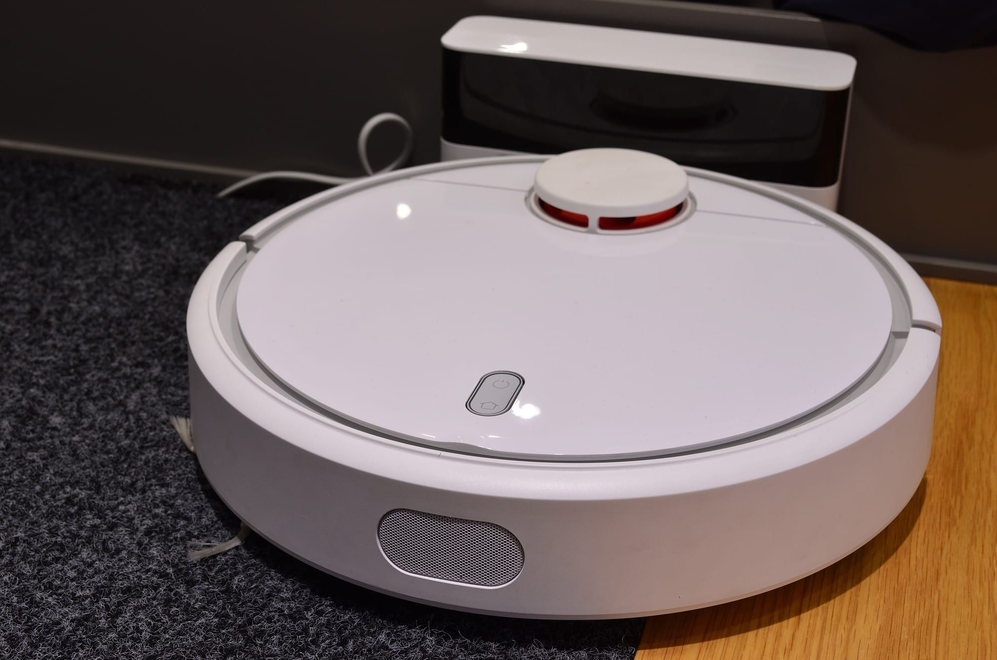 A white robot vacuum cleaner on a wood-effect floor next to its charging dock, with a patch of carpet beside it