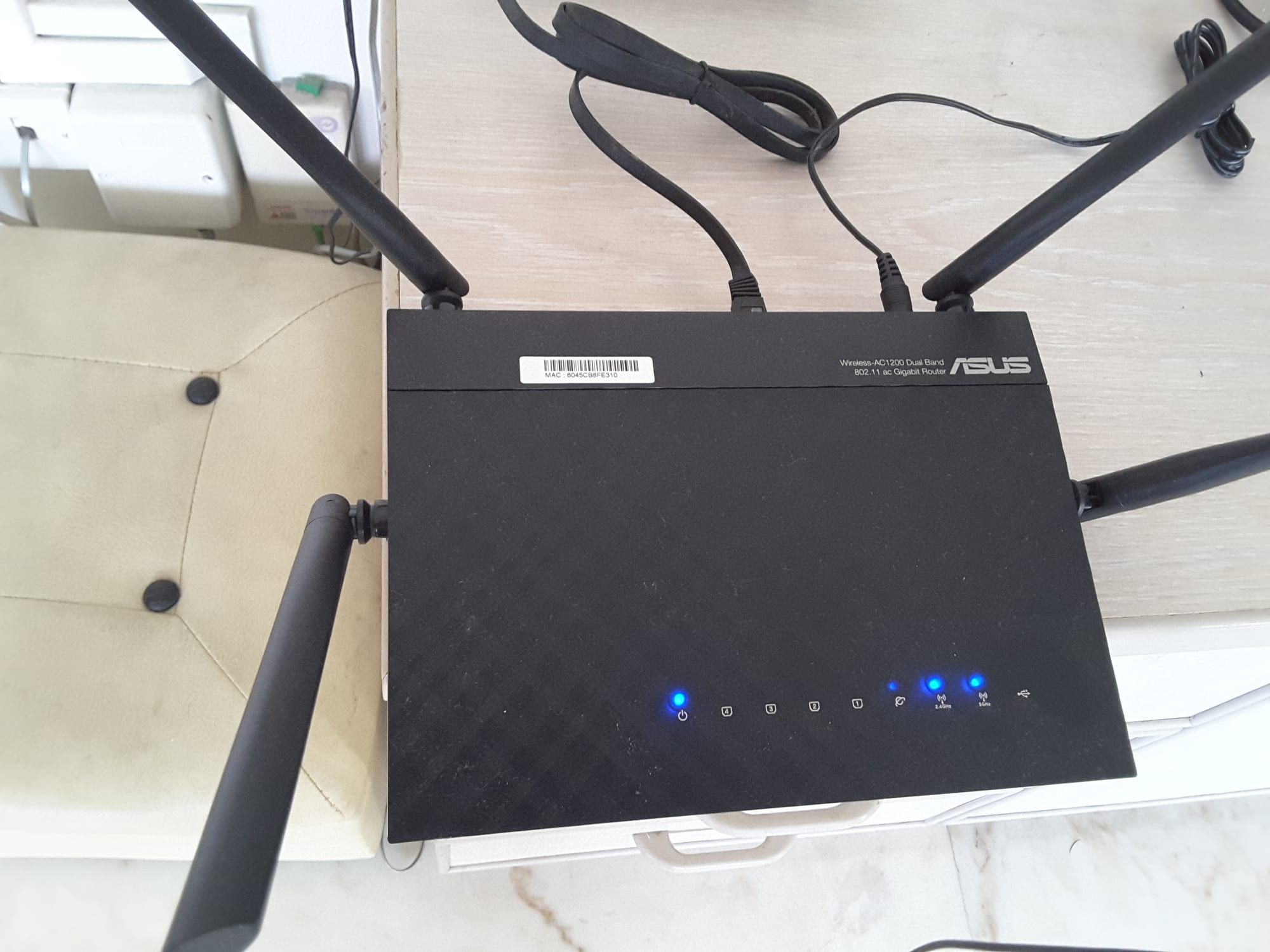 A black Asus RT-AC1200 wireless router with antennas and cables on a table, with blue indicator lights lit