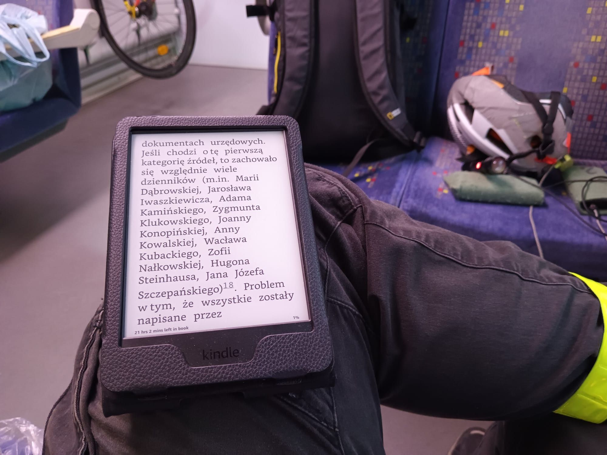 A hand holding an earlier-generation Kindle Paperwhite in a black case, showing a page of Polish text, on a train seat with a backpack and bicycle helmet nearby