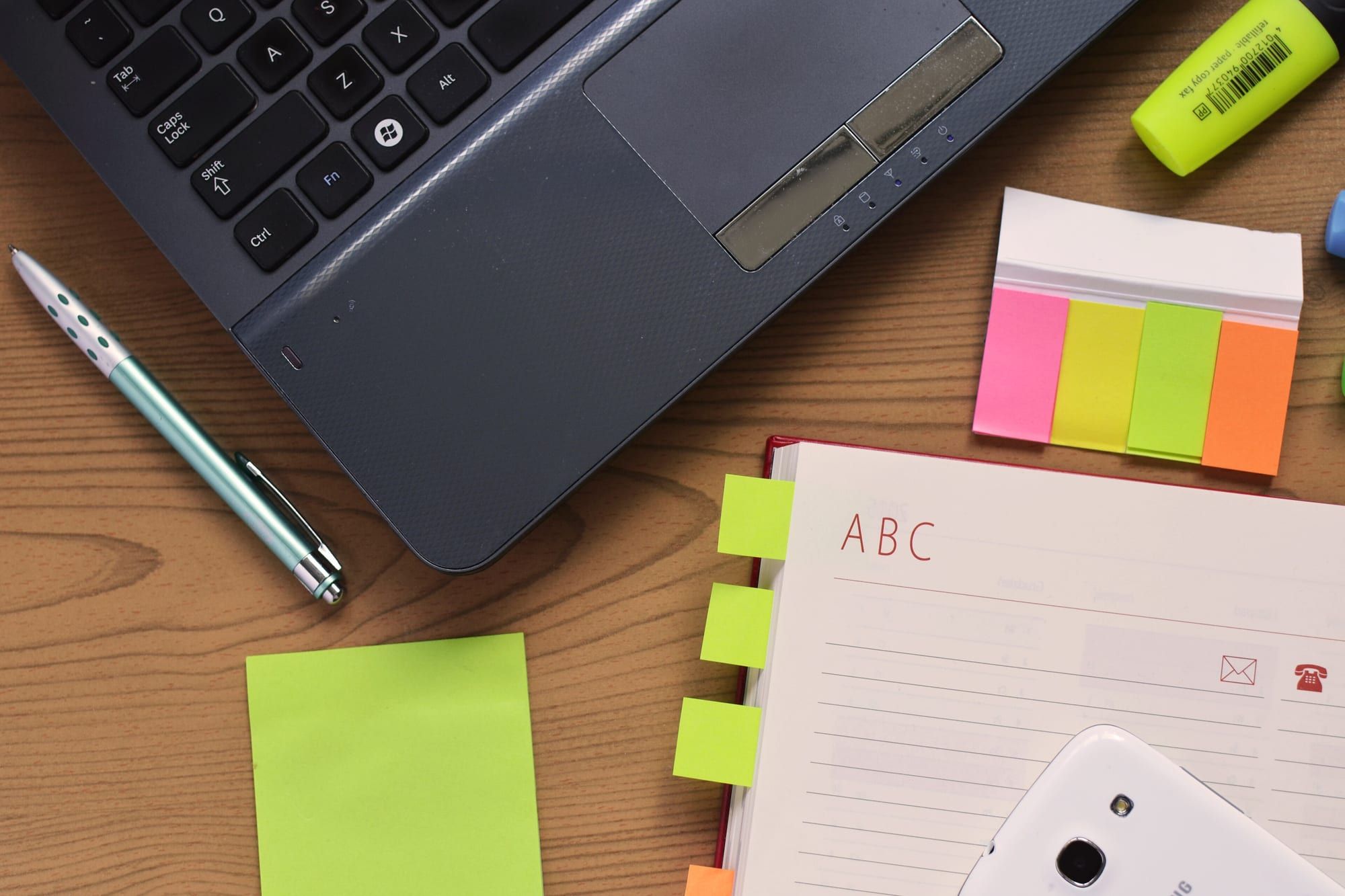 A laptop, notebook, pen and coloured sticky tabs on a wooden desk