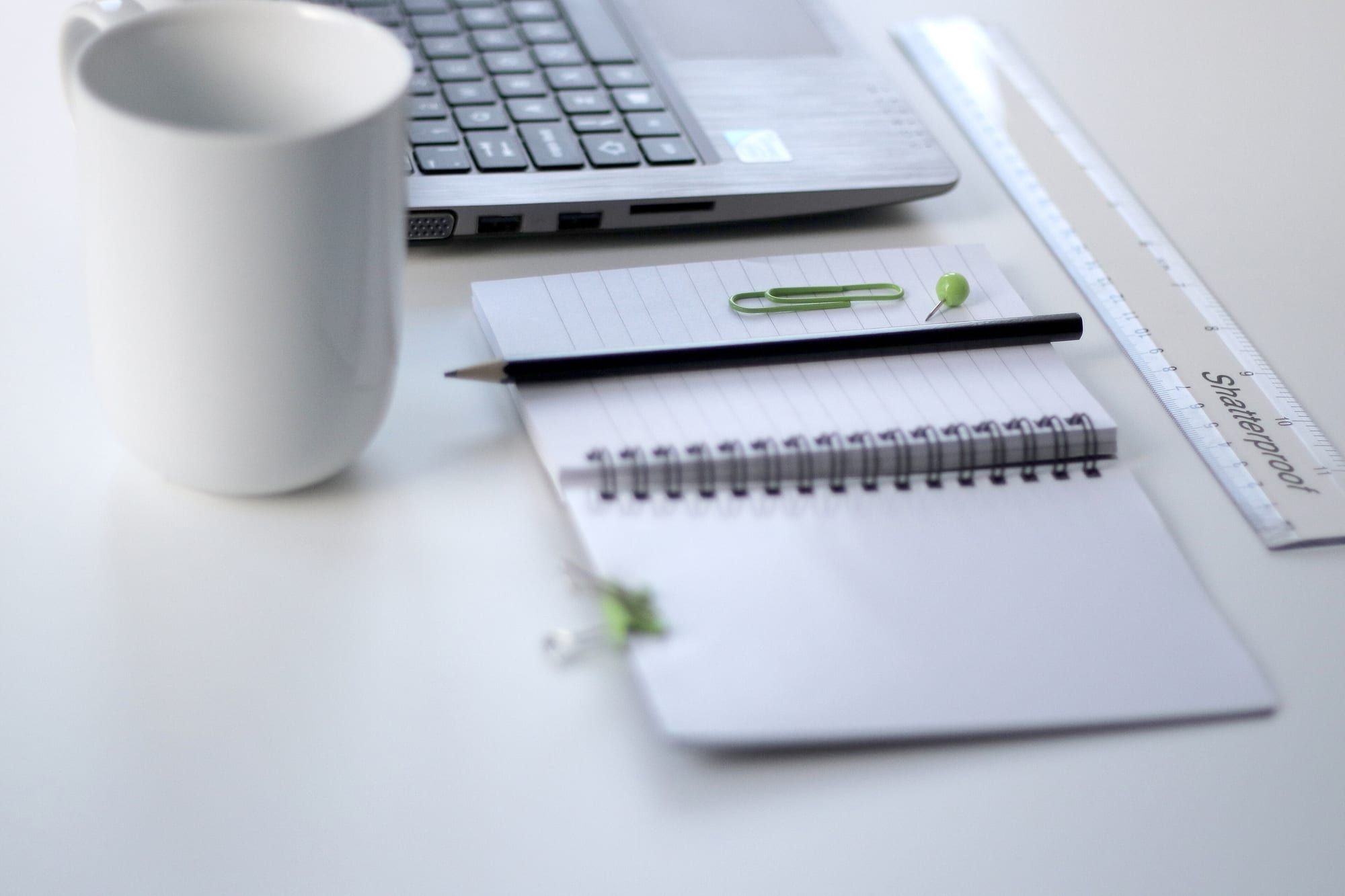A white mug, spiral notepad, pencil, ruler and laptop on a white desk